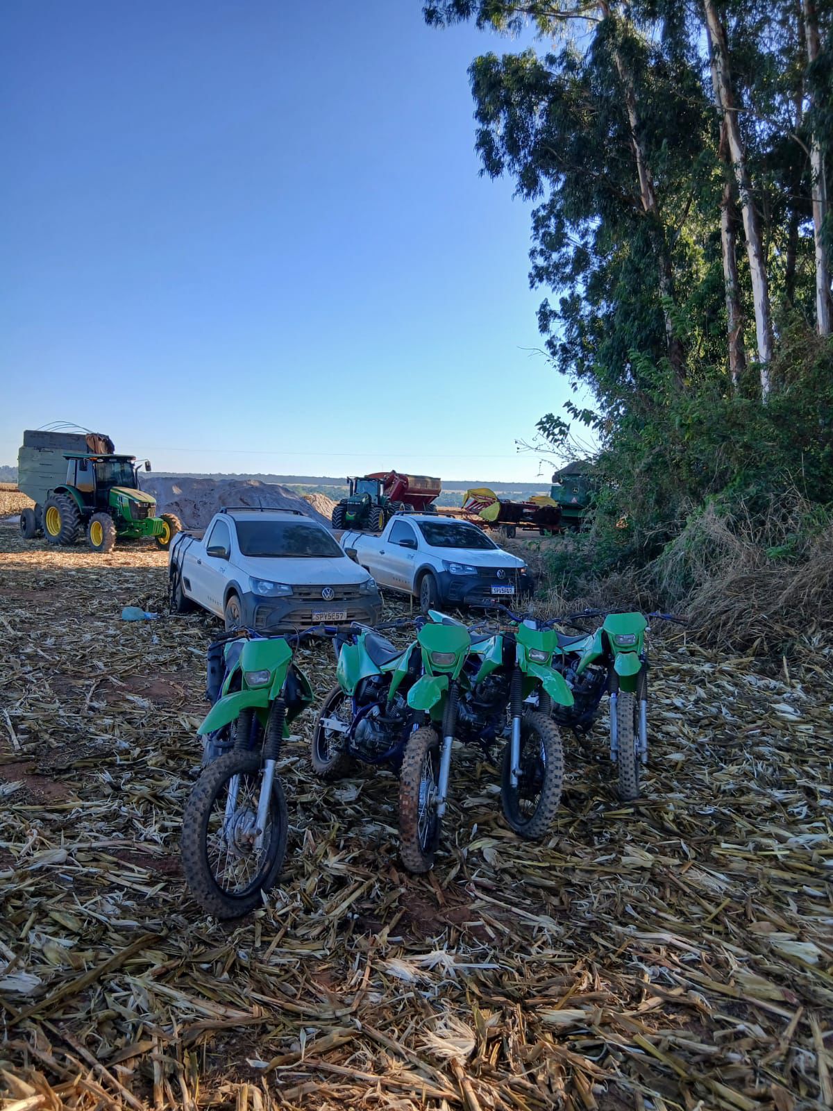 Motocicletas e caminhonetes estacionadas em uma área agrícola, com máquinas ao fundo.